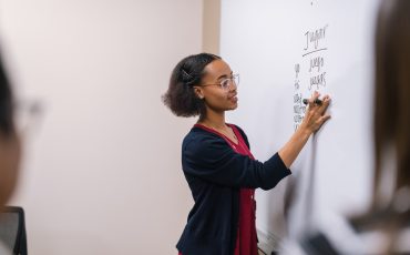 A young ethnic female professor stands at a large whiteboard and writes down Spanish verbs to conjugate.